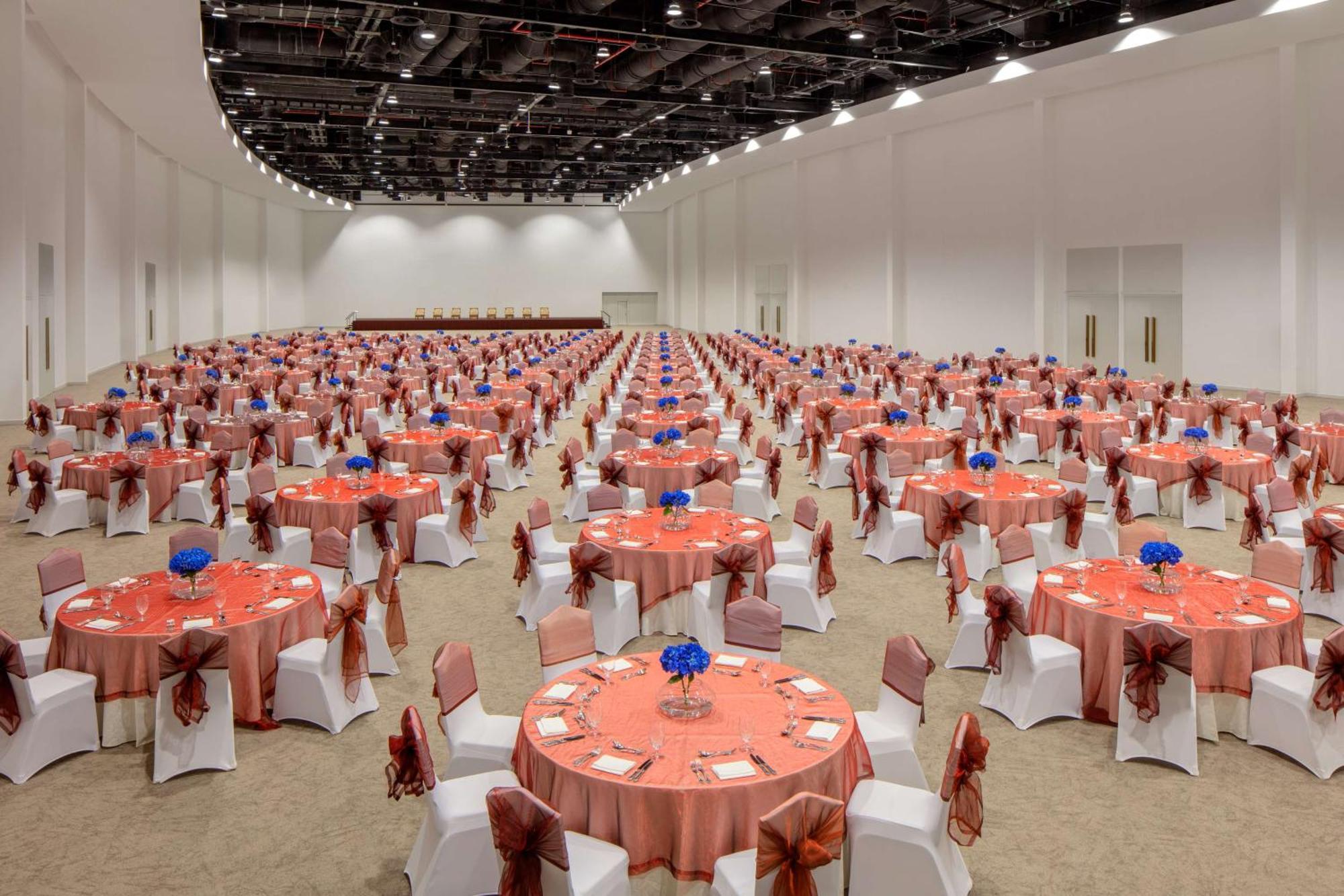 Grand Hyatt Dubai Hotel Ngoại thất bức ảnh The photo shows a large banquet hall set up for an event. There are numerous round tables, each covered with orange tablecloths, and decorated with blue flower arrangements in the center. The chairs are white, adorned with orange sashes. The space ap
