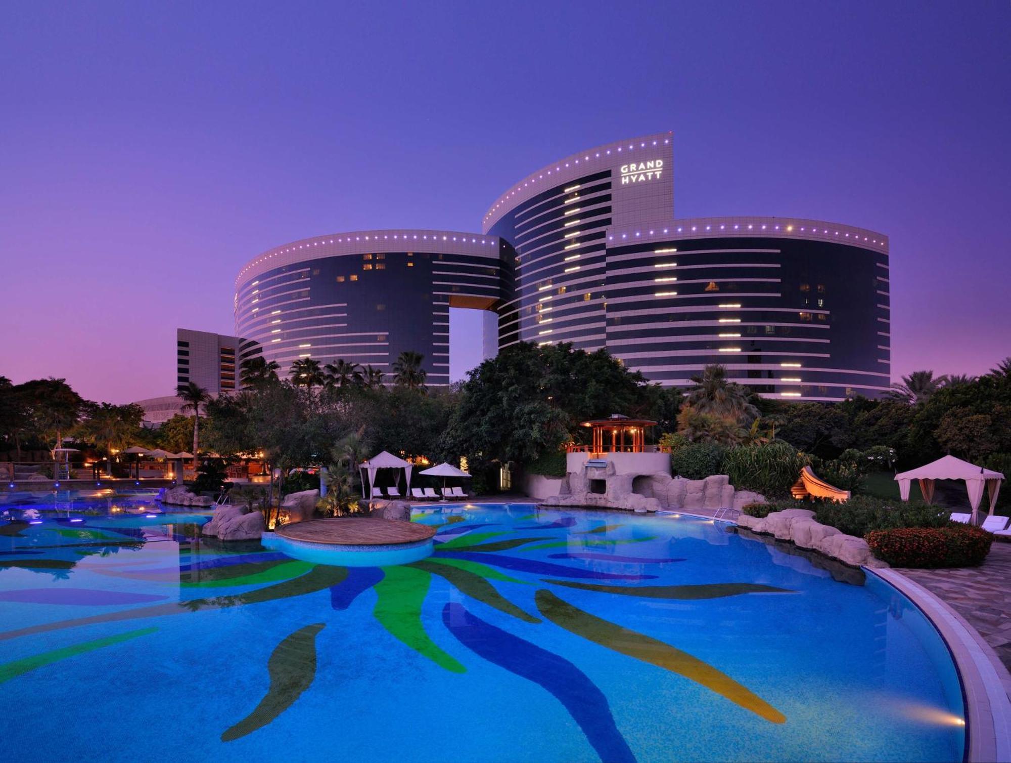 Grand Hyatt Dubai Hotel Ngoại thất bức ảnh The photo shows a luxurious hotel complex set against a twilight sky. The architecture features modern, curved buildings with large windows, reflecting the evening light. In the foreground, there is an expansive pool featuring colorful patterns on th